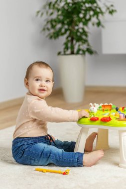 Gülümseyen bebek evde yerde oturuyor, küçük bir aktivite masasında renkli etkileşimli öğrenme oyuncağıyla meşgul oluyor..