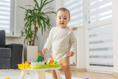 Açık pijamalı bebek, parlak bir oturma odasında renkli bir oyuncak masasının yanında duruyor, oyun oynayarak keşfediyor ve öğreniyor..