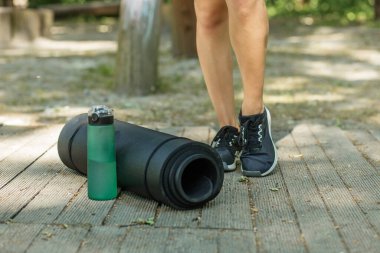 Açık havada spor aletlerinin yakın çekimi: siyah yoga minderi ve yeşil su şişesi, tahta zeminde duran spor ayakkabılı kadının yanında..
