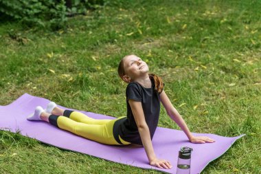 Ergen kız, kobra yogasında çimenli bir parkta mindere uzanıp dinlenmenin ve dışarıda sağlıklı olmanın tadını çıkarıyor..