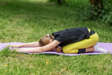 Genç kız, dinlenme, farkındalık ve esneklik üzerine odaklanarak, sessiz yeşil bir parkta yoga minderinde poz veriyor.