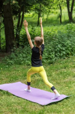 Genç bir kız yoga antrenmanı sırasında yeşil orman parkında huzurlu bir minder üzerinde savaşçı pozu veriyor..