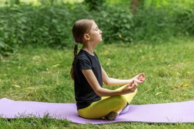 Genç bir kız nilüfer çiçeğinin içinde gözleri kapalı oturuyor, yeşil park ortamında yoga minderinde huzur içinde meditasyon yapıyor..