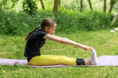 Ergen kız yeşil bir parkta yoga minderinde bacak esnetiyor, esneklik ve farkındalığa odaklanıyor..