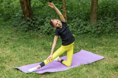 Mutlu genç kız, yemyeşil bir parkta paspasın üzerinde yoga yapıyor, esneklik ve sağlığını arttırıyor..
