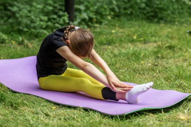 Odaklanmış genç kız, çimenli bir parkta yoga minderinde otururken ayak parmaklarına dokunmak için öne doğru uzanıyor..