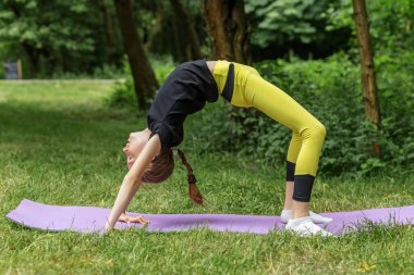 Genç kız ormandaki yoga minderinin üzerinde derin bir ters dönüş ya da tekerlek pozu veriyor. Açık havada güç ve esneklik gösteriyor..