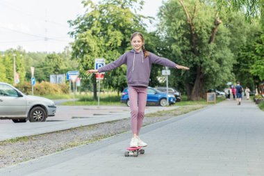 Gülümseyen kız, şehir parkında kaldırımda yürürken kaykayın üstünde denge kuruyor, açık havada aktif bir günün tadını çıkarıyor..
