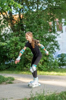 Koruyucu vitesli patenlerle gülümseyen genç bir kız güneşli bir günde yeşil bir yolda güvenle yürür..