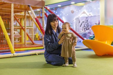Anne, renkli bir oyun merkezinde gülümseyen bir bebekle diz çöküp aile ve çocukluk zamanlarının keyfini çıkarıyor..