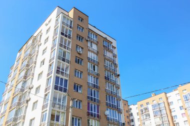 Şehir merkezinde canlı mavi gökyüzüne karşı inşa edilmiş, balkonları ve cam pencereleri olan modern yüksek katlı bir apartman kompleksi..