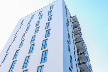 Temiz beyaz duvarları, yerden tavana pencereleri ve açık mavi gökyüzünün altında siyah metal balkonları olan modern yüksek binalar..