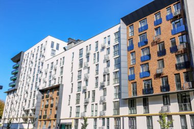 Modern mimari, beyaz ve tuğla ön cephe, dikey pencereler ve mavi gökyüzünün altında renkli cam balkonlar içeren uzun konutlar..