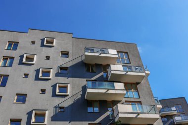 Minimalist gri cephesi, cam balkonları ve mavi gökyüzünün altında kare ve dikdörtgen ahşap çerçeveli pencereleri olan modern konut binaları..