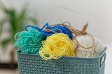 Renkli iplik toplarının yakın plan görüntüsü, desenli plastik bir sepetin içine örgü iğneleri, kapalı bir yere, bulanık arka plan dekoru ile yerleştirilmiş.
