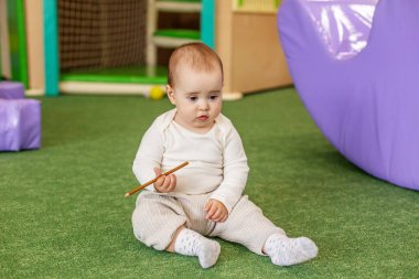 Yumuşak bir oyun odasında elinde bir kalemle oturan sevimli bebek, erken merakı, çocuk gelişimini ve kapalı alanda güvenli oyun ortamını gösteriyor..