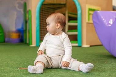 Sevimli bir bebek kreşte yumuşak bir zeminde oturuyor, kapalı oyun saatinde çevreyi keşfediyor, erken gelişim, merak ve çocuk bakımı ortamı gösteriyor..