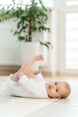 Beyaz tulumlu sevimli bebek yerde uzanıyor, ayaklarıyla oynuyor doğal ışık ve bitkilerle dolu minimalist bir odada..
