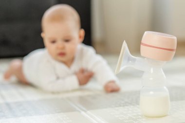 Ön planda taze süt olan elektrikli göğüs pompası, arka planda parlak, temiz, rahat ev içinde yerde uzanan bebek..