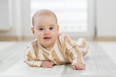 Bej ve beyaz çizgili pijamalarıyla gülümseyen bebek yumuşak zeminde, parlak modern odada doğal ışıkla yatıyor..