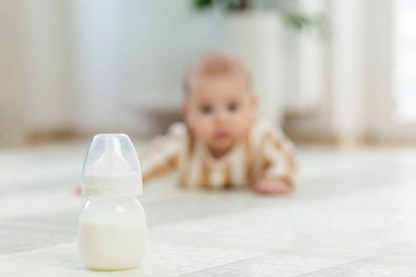 Keskin odaklı süt şişesi, arka planda bulanık çizgili pijamalı emekleyen bir bebek, yumuşak iç mekanda yere uzanıyor..