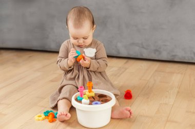 Tahta zeminde oturan sevimli kız bebek renkli şekilli oyuncaklarla oynuyor, erken çocukluk gelişimi ve evde öğrenim aktiviteleriyle uğraşıyor..