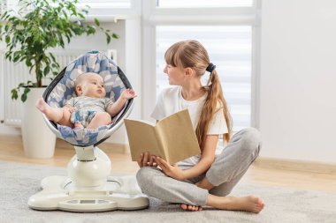 Değer veren abla yerde oturup küçük kardeşine kitap okuyor modern bir oturma odasında sıcak bir aile anını paylaşıyor..