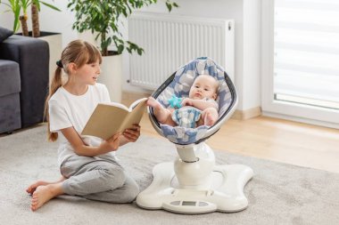 Modern bir oturma odasında yerde oturmuş küçük kardeşine kitap okuyan tatlı bir kız. Aile bağı, çocuk bakımı, erken öğrenim, kardeş ilişkisi, yetiştirme ve ev hayatı kavramı