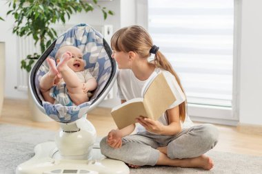Bakımlı abla yerde oturup mutlu küçük kardeşine kitap okuyor. Evde sıcak aile bağları kuruyor..