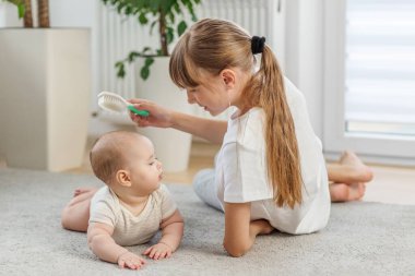 Yumuşak bir halıda uzanmış, bebek kardeşinin saçlarını nazikçe tarayan şefkatli genç bir kız.