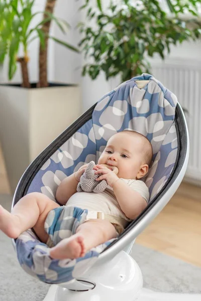 Modern bir bebek koltuğunda uzanmış sevimli bir bebek, parlak ve rahat bir oturma odasında yumuşak bir oyuncağı çiğniyor..