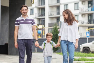 Gülümseyen anne ve baba, genç oğullarını modern bir mahallede okula bırakırken el ele tutuşuyorlar.