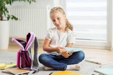 Sırt çantası ve defteriyle yerde oturan genç bir kız, okul konseptine ve ev içi eğitimine geri dönüyor..