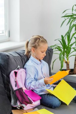 Pembe sırt çantalı genç kız kanepeye oturmuş renkli gazeteler okuyor, okul ödevlerine ve ilköğretim konseptine geri dönüyor..