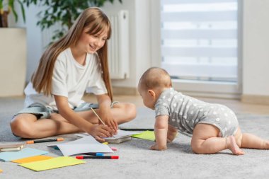 Gülümseyen abla işaretleri kağıda çizerken küçük kardeş ona doğru sürünüyor, evde yaratıcı aile zamanlarının tadını çıkarıyor..