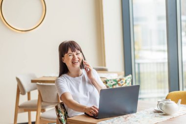 Akıllı telefondan konuşan gülümseyen kadın, bilgisayarını açık kafe içinde kullanıyor, uzaktan çalışmayı, online iletişimi ve modern serbest yaşam tarzını temsil ediyor..