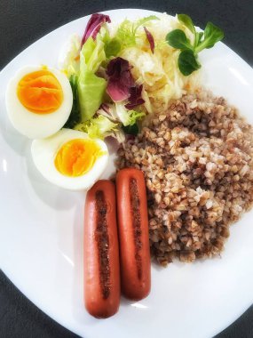 Sağlıklı kahvaltı, öğle yemeği, yemek, ızgara sosis, lahana turşusu, salata, yeşiller, haşlanmış yumurta, yumurta, karabuğday lapası beyaz tabakta.
