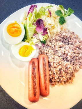 Sağlıklı kahvaltı, öğle yemeği, yemek, ızgara sosis, lahana turşusu, salata, yeşiller, haşlanmış yumurta, yumurta, karabuğday lapası beyaz tabakta.