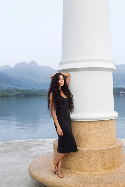 Bella donna in abito nero con lunghi capelli neri in piedi vicino al faro — Foto Stock
