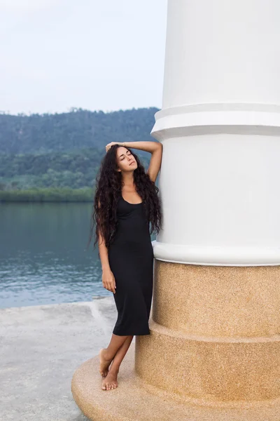 白い壁近くに立っている長い黒髪と黒のドレスで美しい女性 — ストック写真