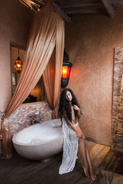 Молодая женщина в белом белье расслабляется возле каменной ванны, полной пены, в красивой роскошной ванной комнате — стоковое фото