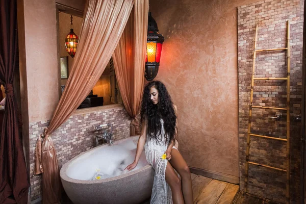 Jeune femme en lingerie blanche relaxante près d'un bain en pierre pleine de mousse dans une belle salle de bain de luxe — Photo