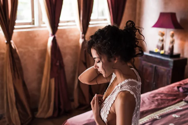 Mujer de pelo negro largo en lencería de seda blanca sentada en la cama en un hotel de lujo por la mañana —  Fotos de Stock