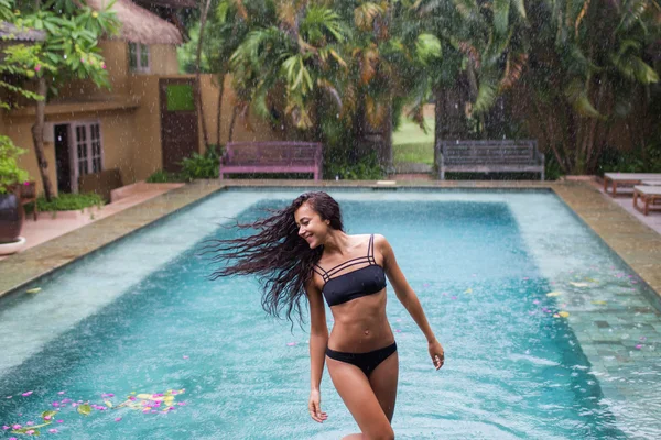 Donna felice godendo in costume da bagno nero sotto la pioggia tropicale vicino alla piscina di lusso — Foto Stock