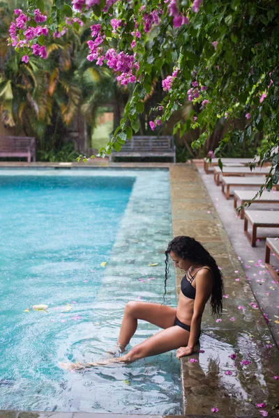 Счастливая женщина в черном купальнике под тропическим дождем возле роскошного бассейна — стоковое фото