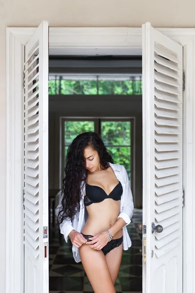 Hermosa mujer con el pelo largo y negro posando cerca de la puerta blanca en lencería trasera. Humor matutino — Foto de Stock