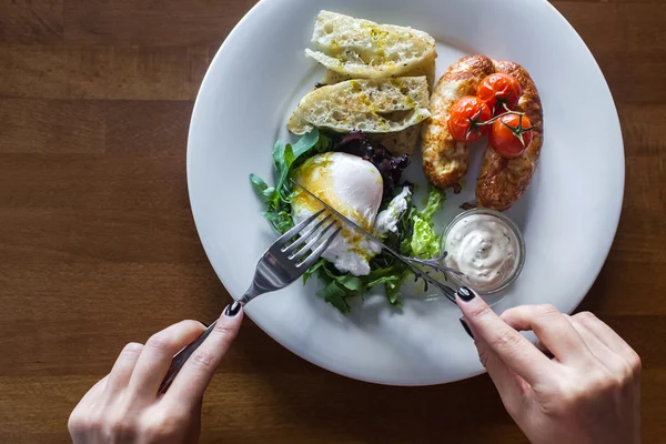 Petit déjeuner avec saucisses frites, tomates cerises cuites au four, œufs pochés et légumes verts. Femme tenant fourchette et couteau — Photo