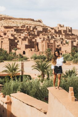 Güzel genç bir kadın arka planda Ait-Ben-Haddou Kasbah manzarasıyla kalenin çitlerinden geçiyor. Fas 'a seyahat. Dikey fotoğraf.