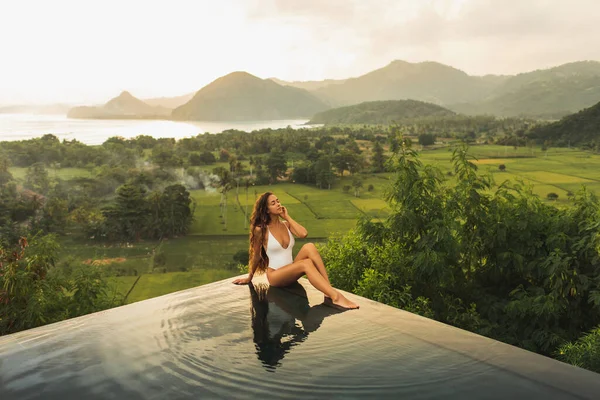 Dream journey in Asia. Woman sitting on edge luxury infinity pool an enjoying amazing mountain, rice terraces and coastline view at sunset. Inspiration travel destination.