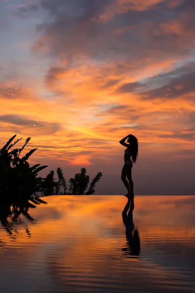 Silhouette of slim woman standing on infinity luxury pool and watching outstanding multicolor sunset.